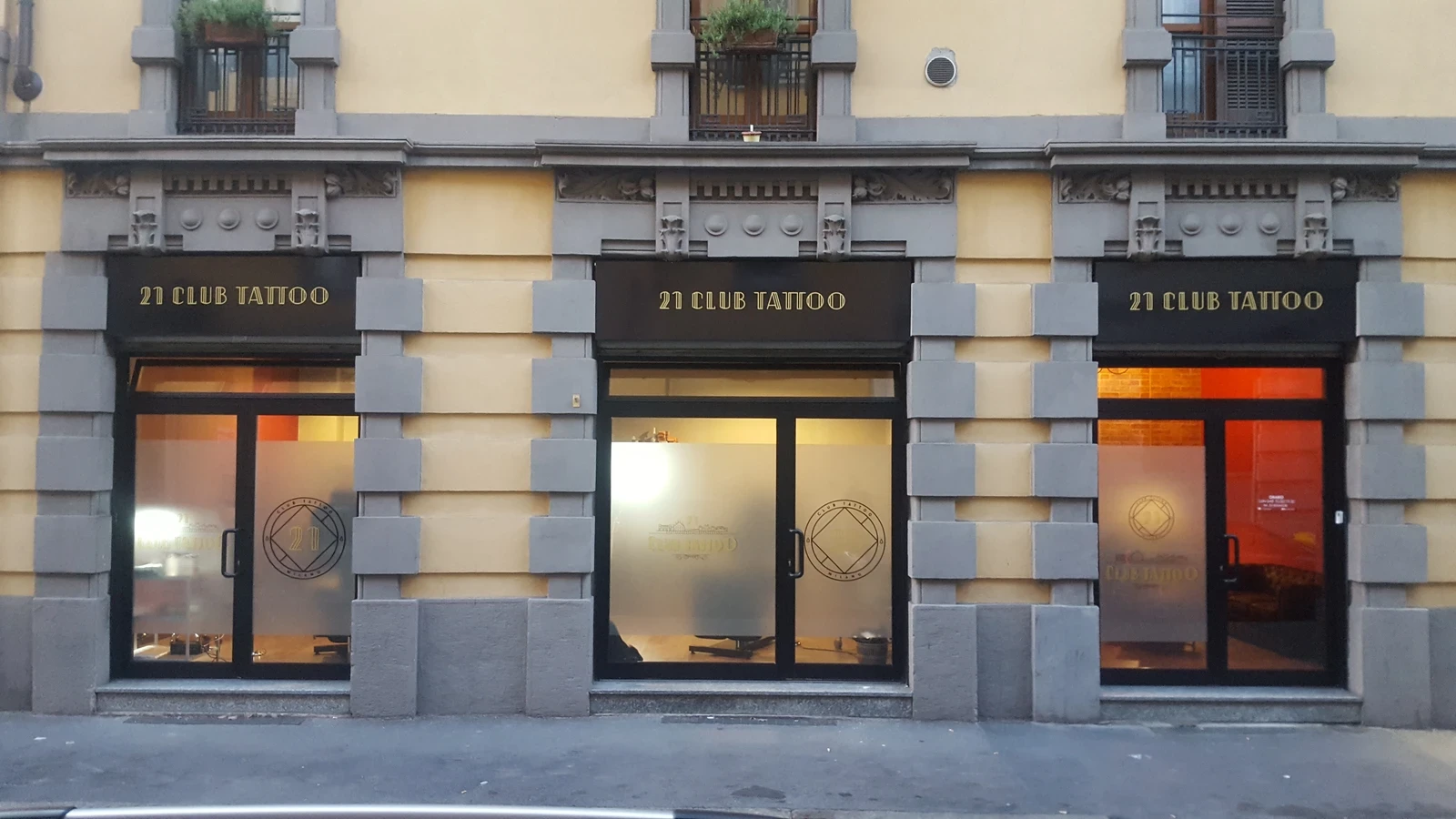Vetrina di uno studio di tatuaggi a Milano, Sailors Tattoo, con tre vetrine luminose e insegne 21 Club Tattoo sull’edificio storico.